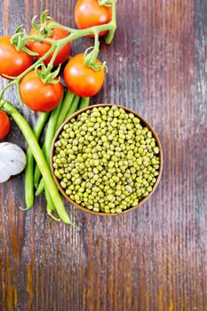 Green lentils mung in a bowl, pods of beans and red tomatoes on the background of a dark wooden board from above