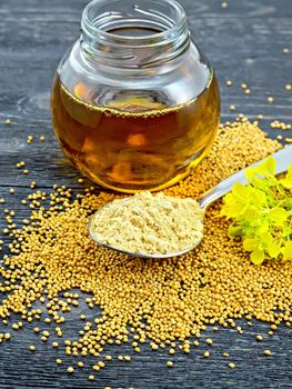 Mustard powder in a metal spoon, oil in a glass jar, seeds and mustard flower on a black wooden board background