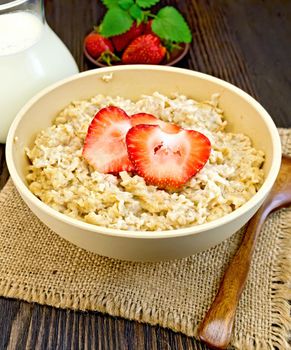 Oatmeal in a bamboo bowl with strawberries on a napkin of burlap, spoon, milk in a glass jar on the background of dark board