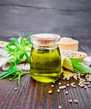 Hemp oil in a glass jar, grain in a spoon and flour in a bowl on sackcloth, cannabis leaves and stalks against a dark wooden board