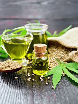 Hemp oil in two glass jars and sauceboat with grain in a sack, a spoon with flour, leaves and stalks of cannabis on a dark wooden board background