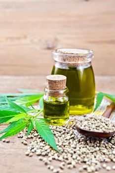 Hemp oil in two glass jars, a spoon with grains on a napkin of burlap, leaves and stalks of cannabis on a wooden board background