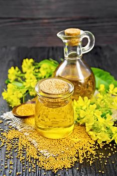 Mustard oil in a glass jar and decanter, mustard seeds on burlap, flowers and leaves on dark wooden board