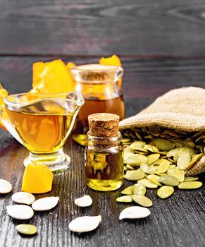 Pumpkin oil in a glass vial, gravy boat and a jar, seeds in a bag and on table, slices of vegetable on wooden board background