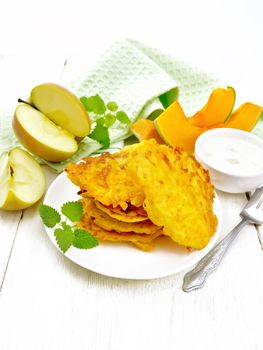 Pumpkin and apple fritters in a plate, sour cream in a bowl, napkin, mint and fork on wooden board background