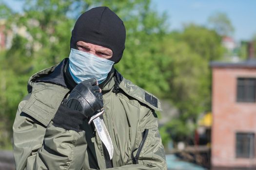 Pensive criminal with a knife in a medical mask and balaclava squints his eyes because of the bright sun