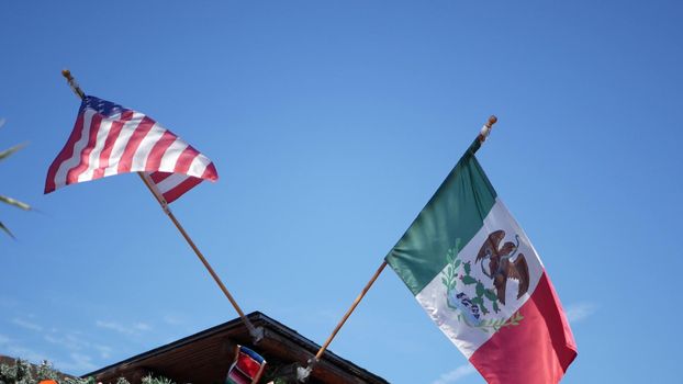 Mexican tricolor and American flag waving on wind. Two national icons of Mexico and United States against sky, San Diego, California, USA. Political symbol of border, relationship and togetherness.