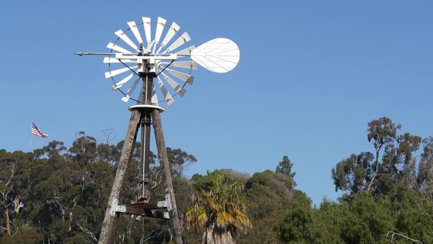 Classic retro windmill, bladed rotor and USA flag against blue sky. Vintage water pump wind turbine, power generator on livestock ranch or agricultural farm. Rural symbol of wild west, rustic suburb.