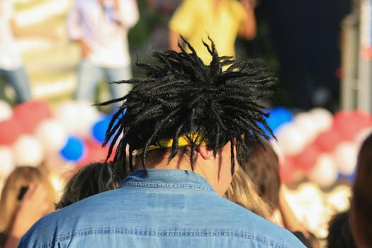 A man in a black dreadlock wig at a street concert. Stands with his back, looks to the side. Sun, summer.