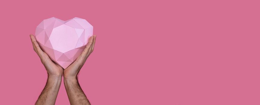 Polygonal three-dimensional pink heart made of paper in the hands of a man. On a pink background. Banner. Copy space. 