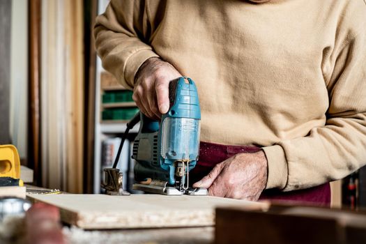 Carpenter's hands with electric jigsaw close-up. Work in a carpentry workshop. A man cuts plywood with an electric jigsaw. Electric tool for woodworking.