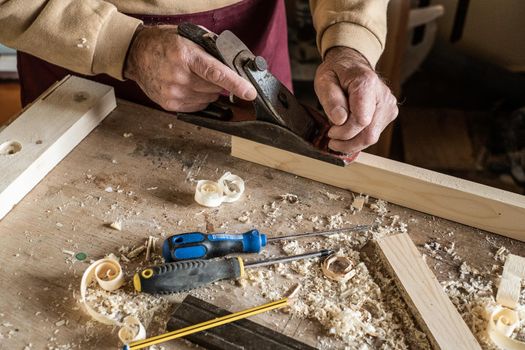 Carpenter man scraping curled wood scraps with hand plane tool and wooden plank.