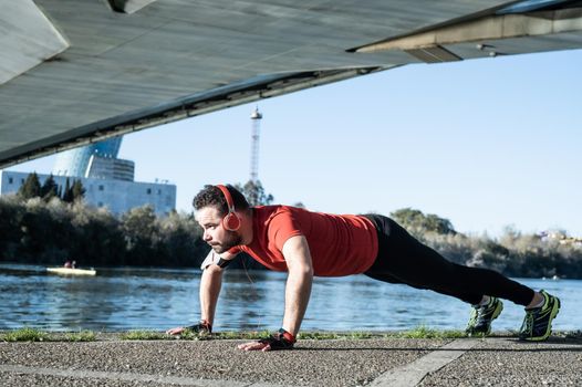 young man doing push-ups with a red t-shirt and music headphones, in an urban environment. healthy living concept