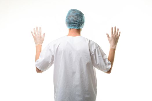 A male doctor stands with his hands raised up, gloves are on, a mask and a hat are on his head, rear view