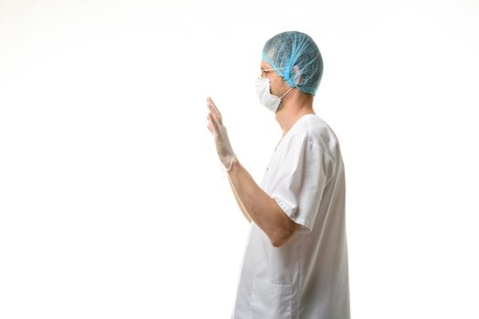 A male doctor stands with his hands raised up, gloves are on, a mask and a hat are on his head, side view