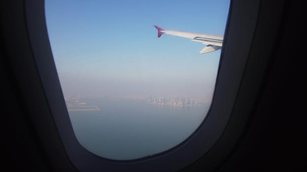 View through an airplane window on the city Qatar. Airplane window view showing wing of a plane flying over city. Travel concept.