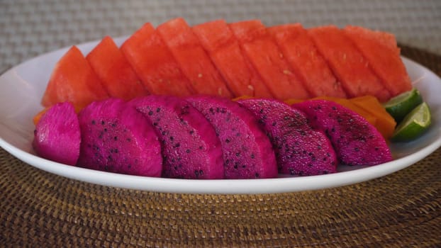 Female hands put on the table Assorted fresh ripe tropical fruits on a plate including Dragon fruit,pitaya, watermelon. Fresh served fruits salad on white dish.