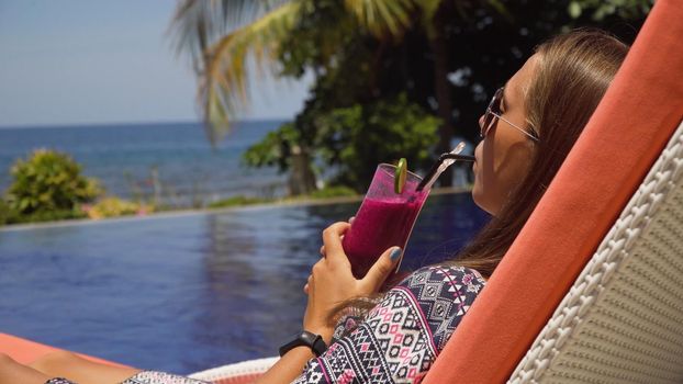 Beautiful young girl with glass of cocktail relaxing near swimming pool. Girl in sunglasses with cocktail in seafront near the bassin amongst the palm trees on the background of the sea. Travel, vacation concept.