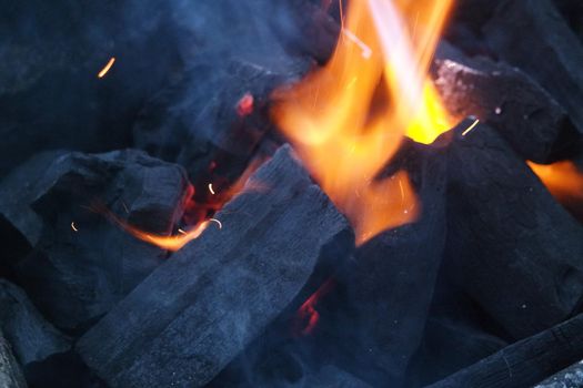 Fire on logs in fire pot with embers and burning coal. Bonfire with blazing red and orange fire flames. Fire background with woods and planks on fire at night view.