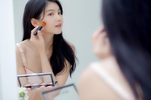 Beauty young asian woman happy and smile with face looking mirror applying makeup with brusher of cheek in the room, beautiful of girl holding brush make-up on facial, skin care and cosmetic concept.