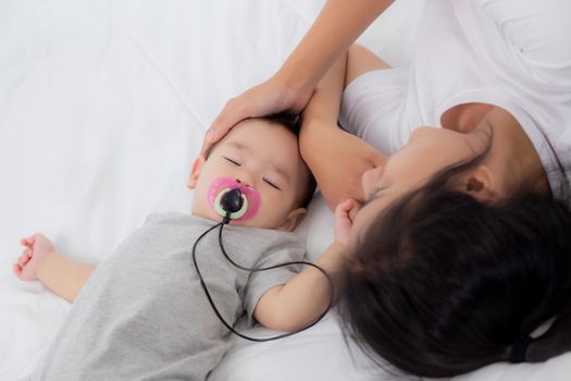 Young asian mother touch head of little baby girl with tender on bed in the bedroom, mom love newborn and care, woman with expression with child together, parent and daughter, family concept.