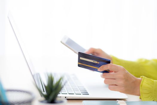 Closeup young asian business woman using smart phone and holding credit card while online shopping and payment with laptop computer on desk at home, female holding debit card, communication concept.