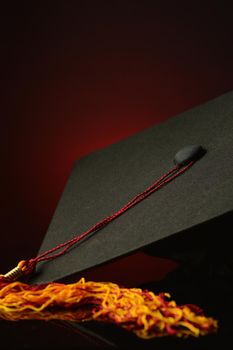 A closeup vertical shot of a mortarboard grad hat over a dark red and black background.