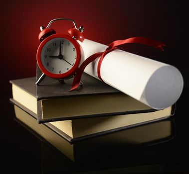 A diploma on top of a couple books shows the successful payoff of school studies while an alarm clock reminds us of the important timeline at hand.