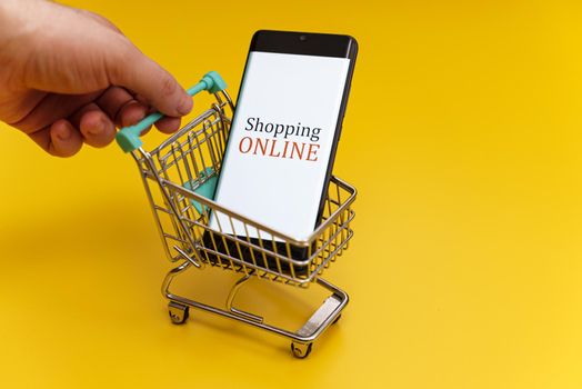 Shopping cart with smartphone on yellow background. Online shopping concept.