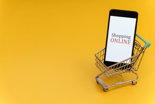 Shopping cart with smartphone on yellow background. Online shopping concept.