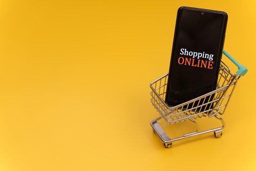 Shopping cart with smartphone on yellow background. Online shopping concept.