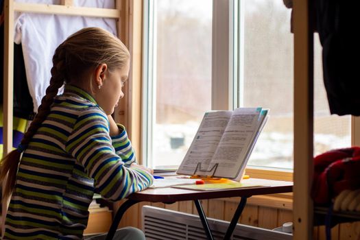 Schoolgirl carefully reads the task in the textbook while doing homework while sitting by the window