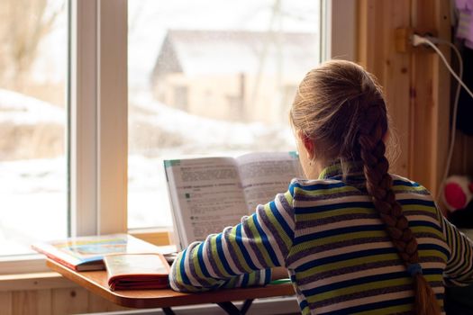 Back view of a schoolgirl sitting by the window and doing homework