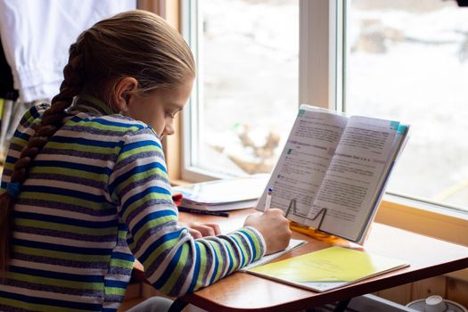 Schoolgirl sits at the table by the window and does homework