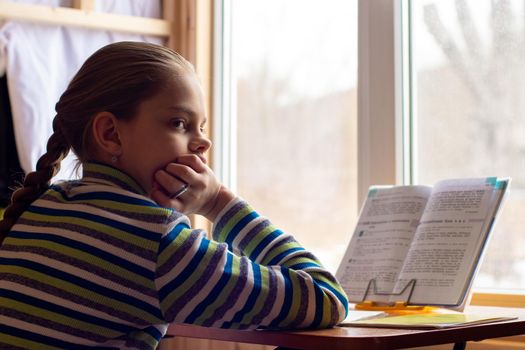 A schoolgirl sits at a table by the window and does her homework, the girl was distracted in thought and looked towards the frame