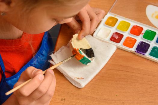 A girl paints a craft from salt dough with watercolors, top view