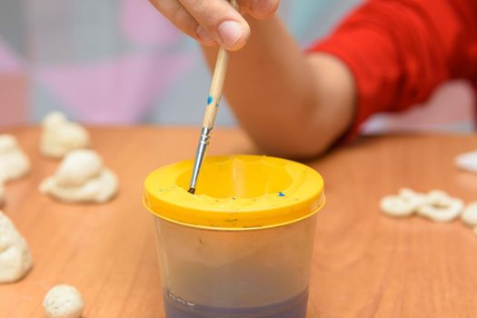 A girl dips a brush into a glass of water painting figures from salt dough