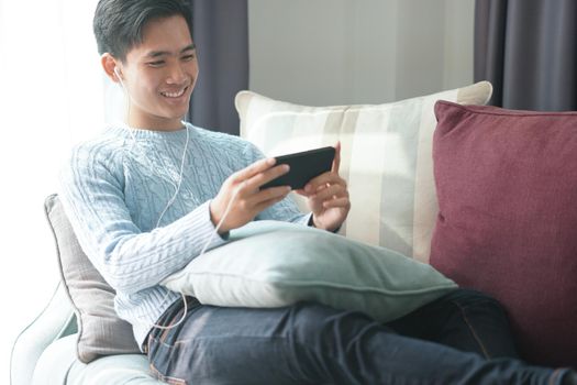 young asian man wearing earphones listening to music on smart phone relaxing on sofa at home