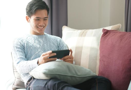 young asian man wearing earphones listening to music on smart phone relaxing on sofa at home
