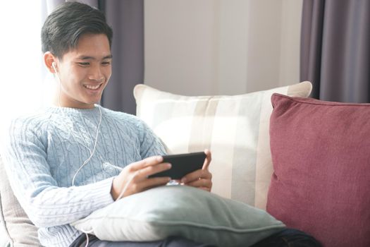 young asian man wearing earphones listening to music on smart phone relaxing on sofa at home