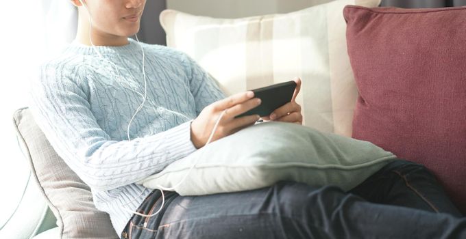 young asian man wearing earphones listening to music on smart phone relaxing on sofa at home