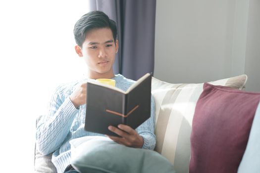 young asian man reading book relaxing on sofa at home. leisure lifestyle