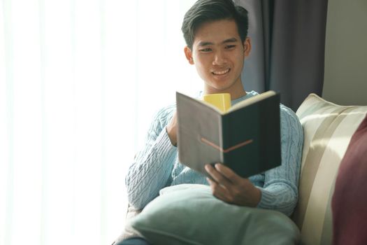 young asian man reading book relaxing on sofa at home. leisure lifestyle