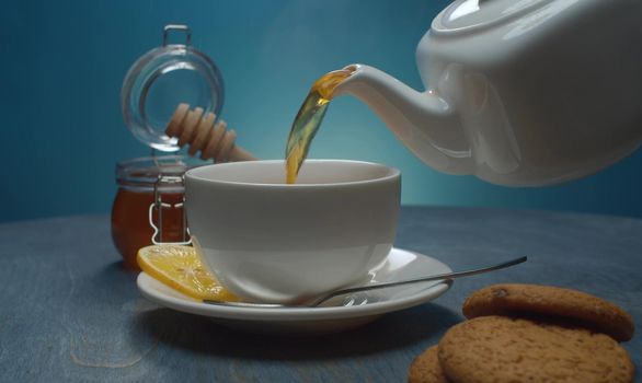 Close-up hot tea pouring into the cup. Honey in glass jar, lemon and cookies on the table