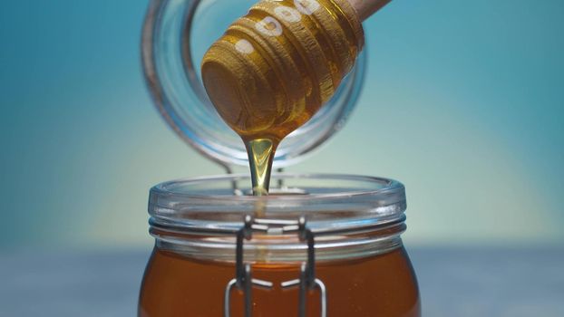 Extreme close up fresh honey pouring from honey spoon into a jar on blue background. Healthy fresh food concept. Camera flying around it. Macro
