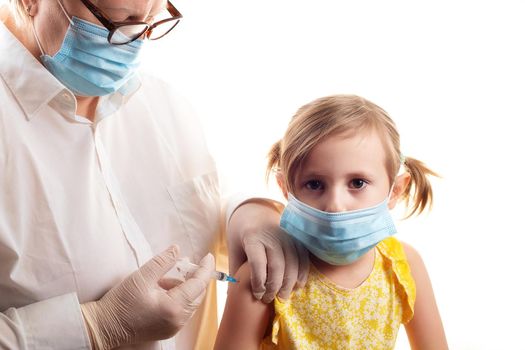 Vaccination concept in the era of coronavirus. Woman Doctor vaccinating cute little girl wearing yellow dress and facial protective mask on white background.