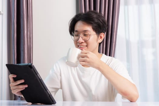 Happy Asian young business handsome man work from home office wear glasses, t-shirt comfortable he smiling and using a black modern smart digital tablet computer to read email or learning on desk