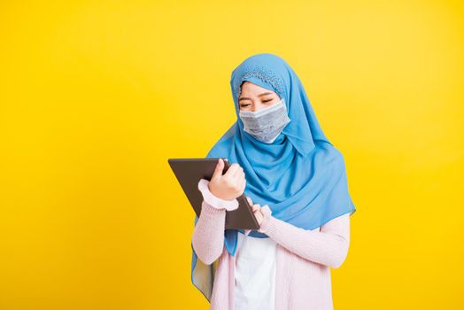 Asian Muslim Arab, Portrait of happy beautiful young woman Islam religious wear veil hijab and face mask protect she quarantines disease coronavirus touch on tablet screen isolated yellow background