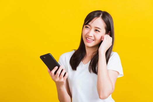 Portrait Asian of a happy beautiful young woman holding a mobile smart phone and wearing wireless headphones listening to music from smartphone studio shot isolated on yellow background