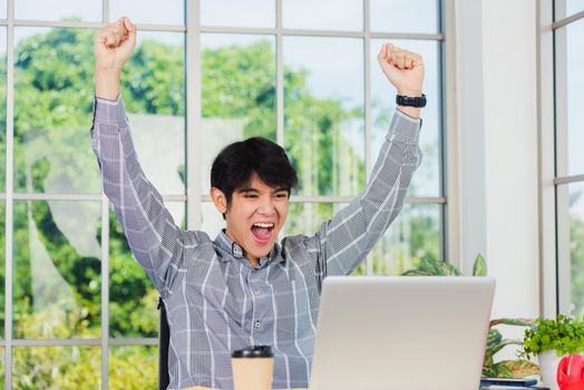 Yes! Happy excited Asian man raising his arm up to celebrate celebrating success. Young businessman using laptop computer at office desk he glad to receive good winner profits from the job